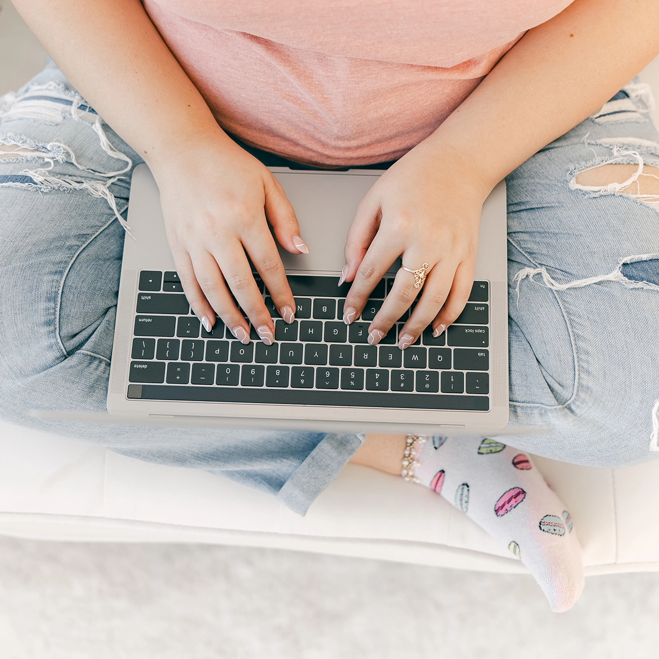 woman in ripped jeans and pink shirt sitting cross-legged, typing on laptop